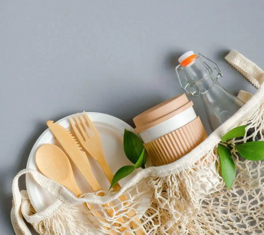 Beige mesh reusable shopping bag holding wooden utensils, a white plate, and eco-friendly glassware with bamboo lid.